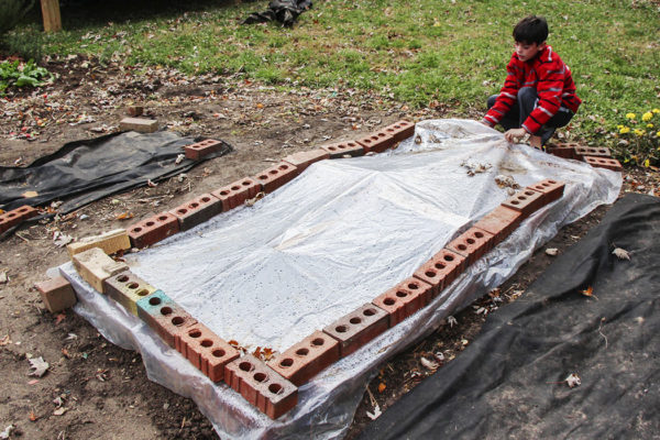A basic cold frame being held shut by bricks with a boy lifting the plastic cover